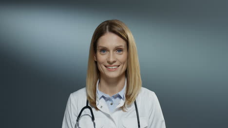 Pretty-young-Caucasian-female-medic-in-white-gown-and-with-stethoscope-on-her-neck-smiling-joyfully-to-the-camera.-Portrait.